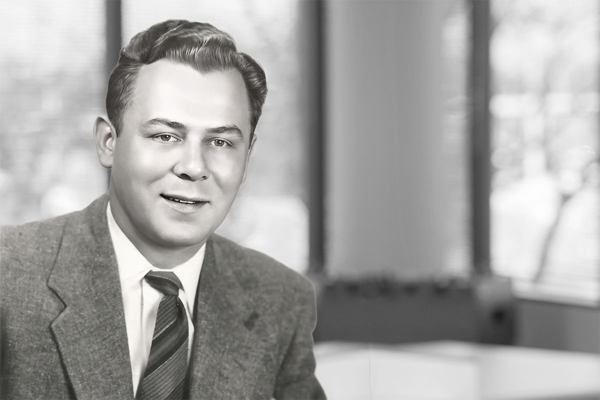 Black-and-white portrait of a clean-cut man wearing a suit and tie, seated indoors with a softly blurred office-style background.