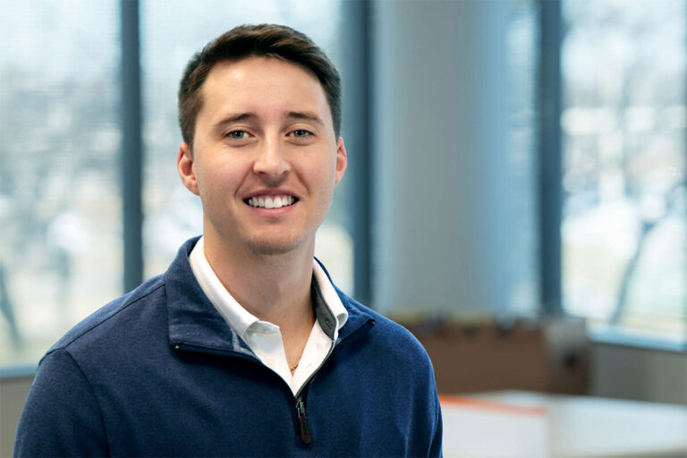 Smiling man with short brown hair wearing a navy quarter-zip pullover over a white collared shirt, standing in a bright office with large windows.