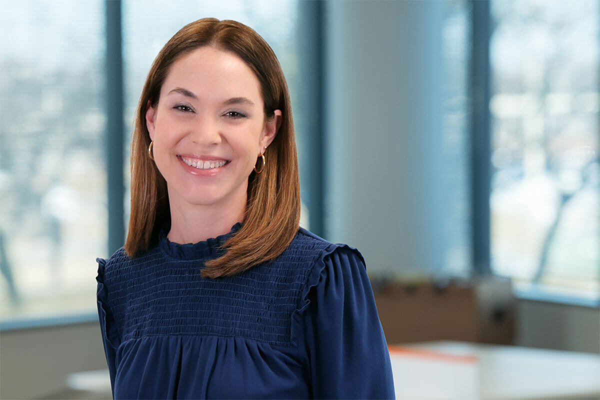 Smiling woman with shoulder-length brown hair wearing a navy blue blouse, standing in a bright office with large windows and a softly blurred background.