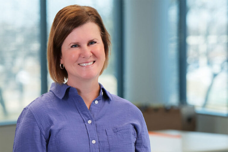 Smiling woman with short brown hair wearing a purple button-up shirt, standing in a bright office with large windows and a softly blurred background.