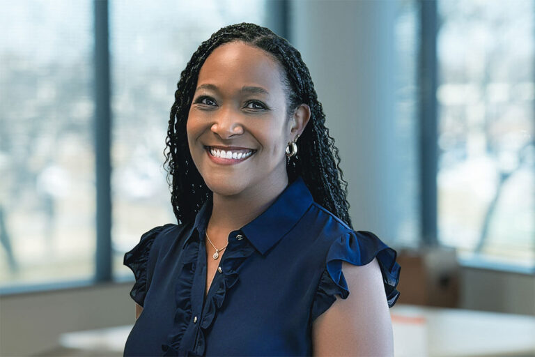 Smiling woman with braided hair wearing a navy blouse with ruffled sleeves, standing in a bright office with large windows and a softly blurred background.