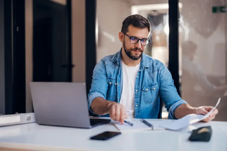 Bearded man wearing glasses and a denim shirt sits at a desk, reviewing paperwork beside a laptop and smartphone in a modern office setting.