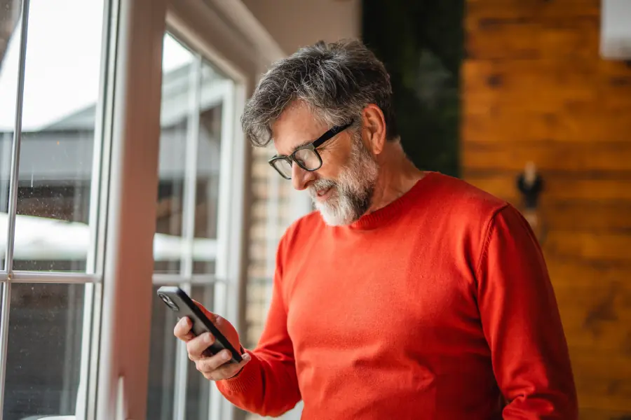 Older man with gray hair and beard wearing glasses and a red sweater, standing by a window indoors while looking down at his smartphone.