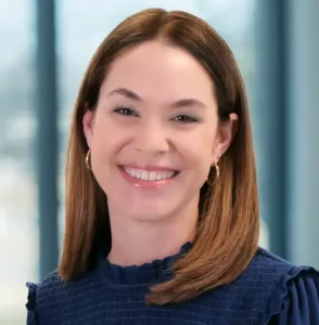 Professional woman smiling in a modern office setting A woman with shoulder-length brown hair smiles at the camera while standing in a bright office with large windows and a softly blurred background.