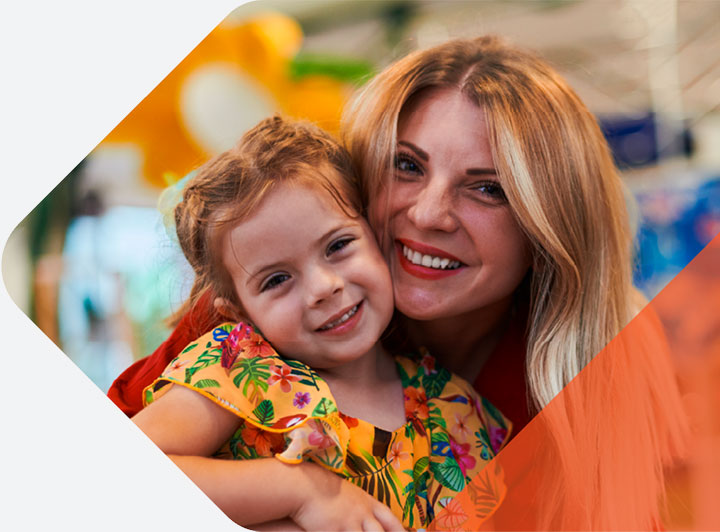 Smiling woman hugs her young daughter as they pose closely together, both wearing colorful outfits in a bright, playful indoor setting.