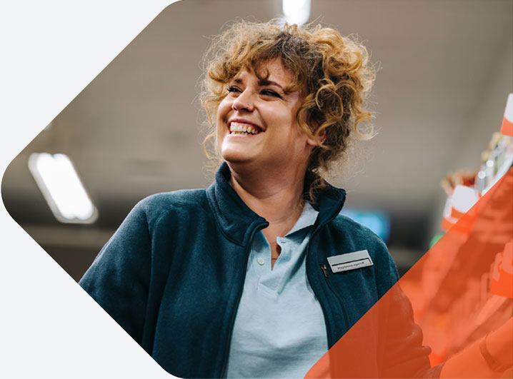 Smiling woman with curly hair wearing a work jacket and name badge stands in a retail aisle, looking off-camera in a bright store environment.