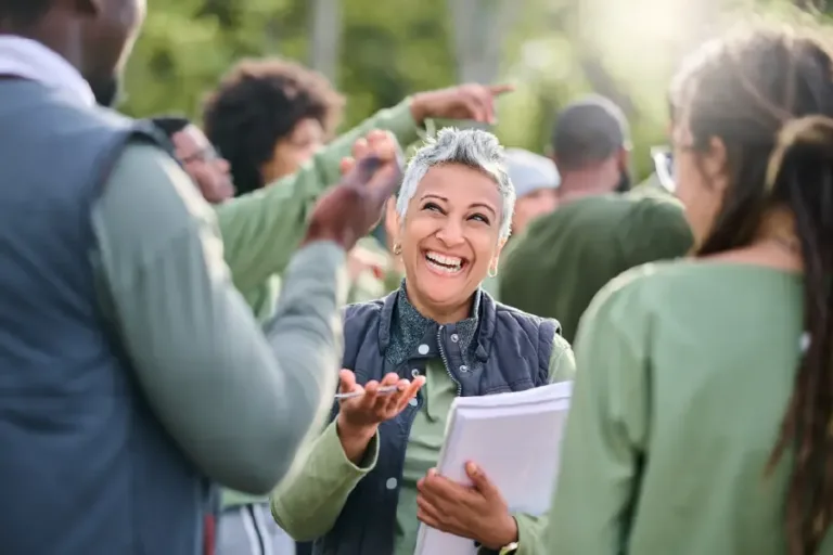 Smiling woman with short gray hair holds papers while laughing and engaging with a diverse group of people during an outdoor team discussion.