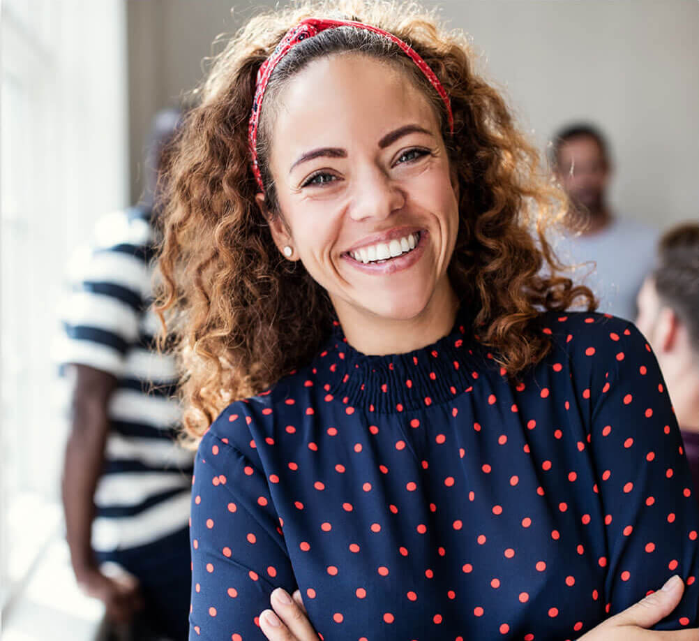 Smiling professional woman standing confidently in a bright office
A woman with curly hair and a red headband smiles with her arms crossed, standing in a modern workspace with colleagues softly blurred in the background.