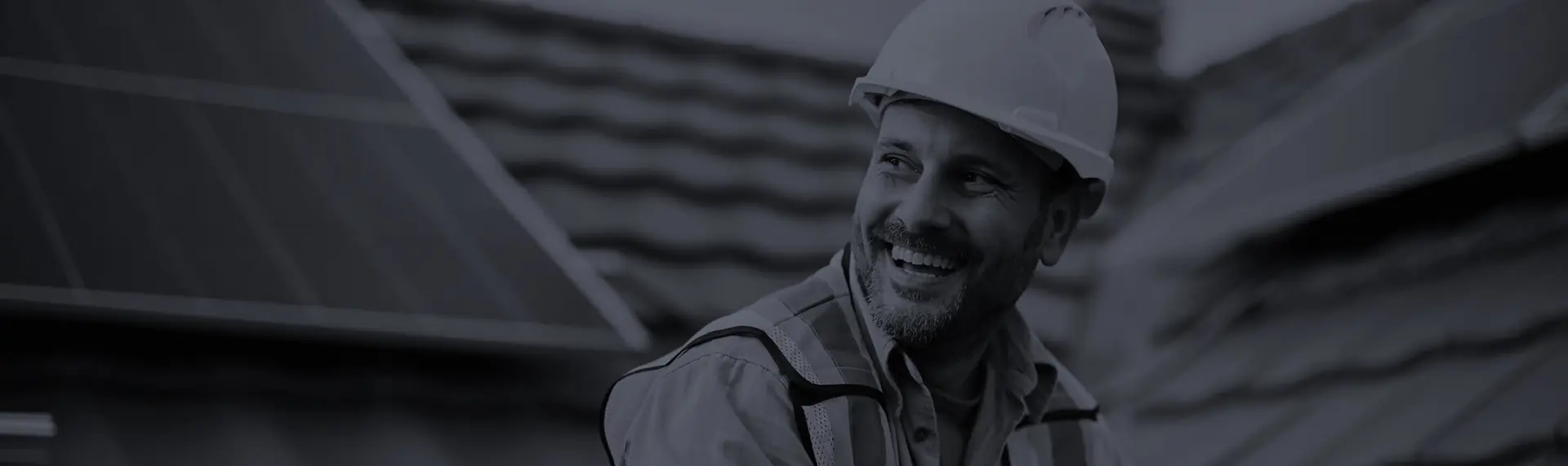 A smiling construction worker wearing a white hard hat and safety vest stands on a rooftop with solar panels and shingles in the background.