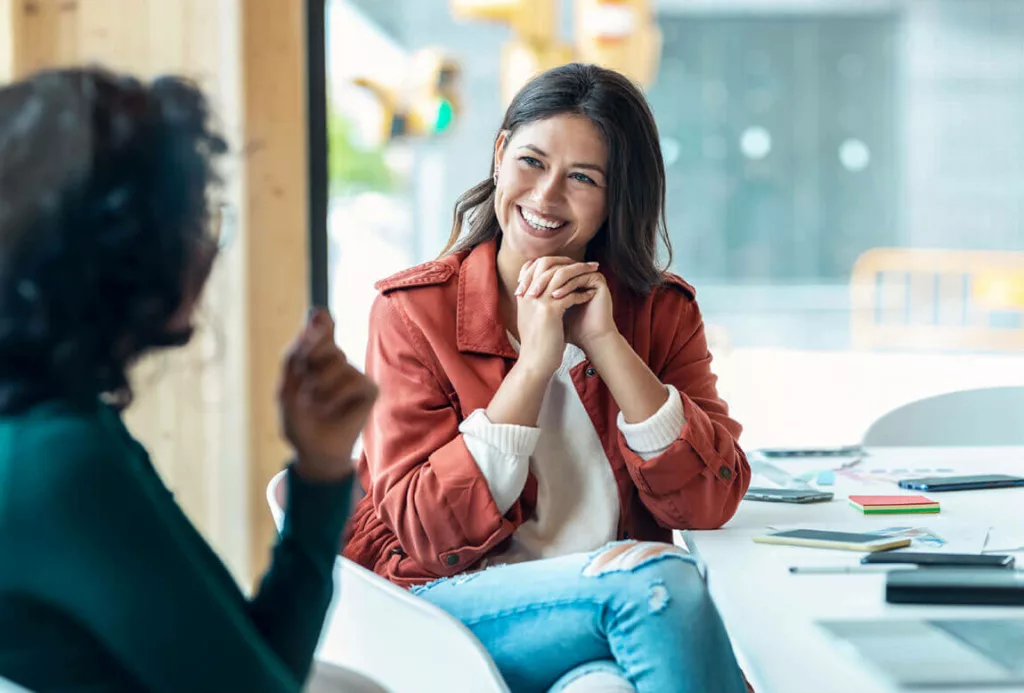 Friendly conversation between two women in a modern office
A woman smiles warmly while sitting at a table and listening to another person speak, with notebooks and a laptop in a bright workspace.