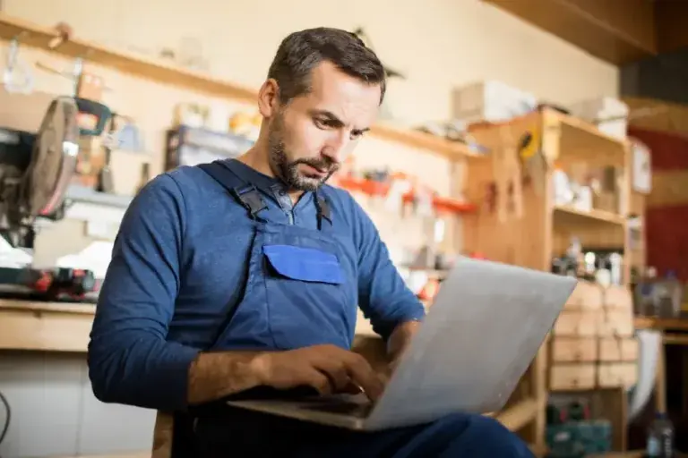 Focused man wearing blue work overalls sits in a workshop, typing on a laptop with tools, shelves, and equipment visible in the background.
