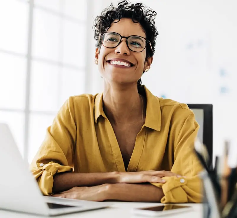 Professional woman smiling at her desk in a bright office A confident woman with short curly hair and glasses smiles while sitting at a desk, with a laptop and office items in a well-lit workspace.