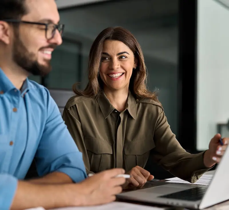 Two colleagues collaborating at a laptop in an office meeting A smiling woman and a man sit side by side at a desk, discussing work while looking at a laptop screen in a modern office setting.