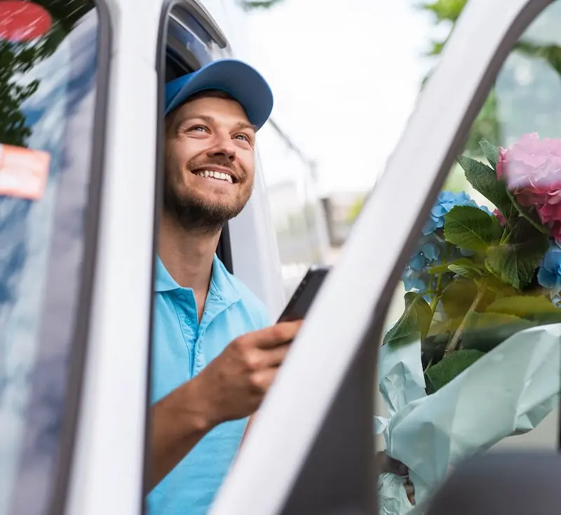 Delivery driver smiling with flowers in a van A cheerful delivery driver sits in a van doorway holding a smartphone, with wrapped bouquets of colorful flowers visible beside him.