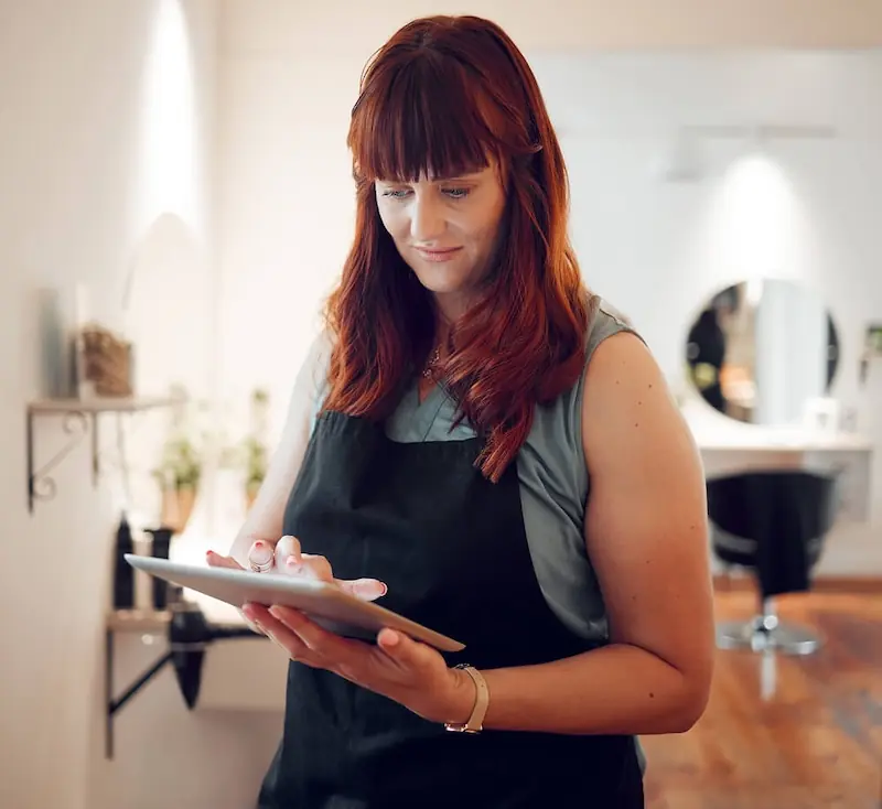Independent professional using a tablet in a studio workspace A woman wearing an apron looks down at a tablet while standing in a bright studio or salon, with mirrors and tools visible in the background.