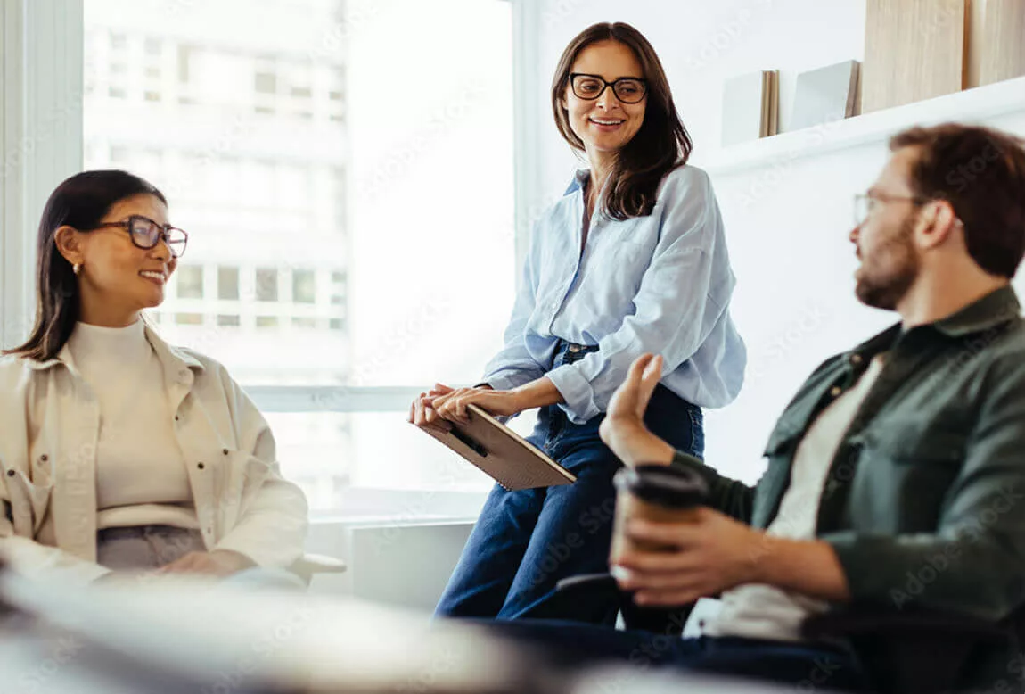Team discussion among colleagues in a bright office setting Three coworkers talk casually in a modern office, with one woman holding a notebook while listening as others share ideas during a relaxed meeting.