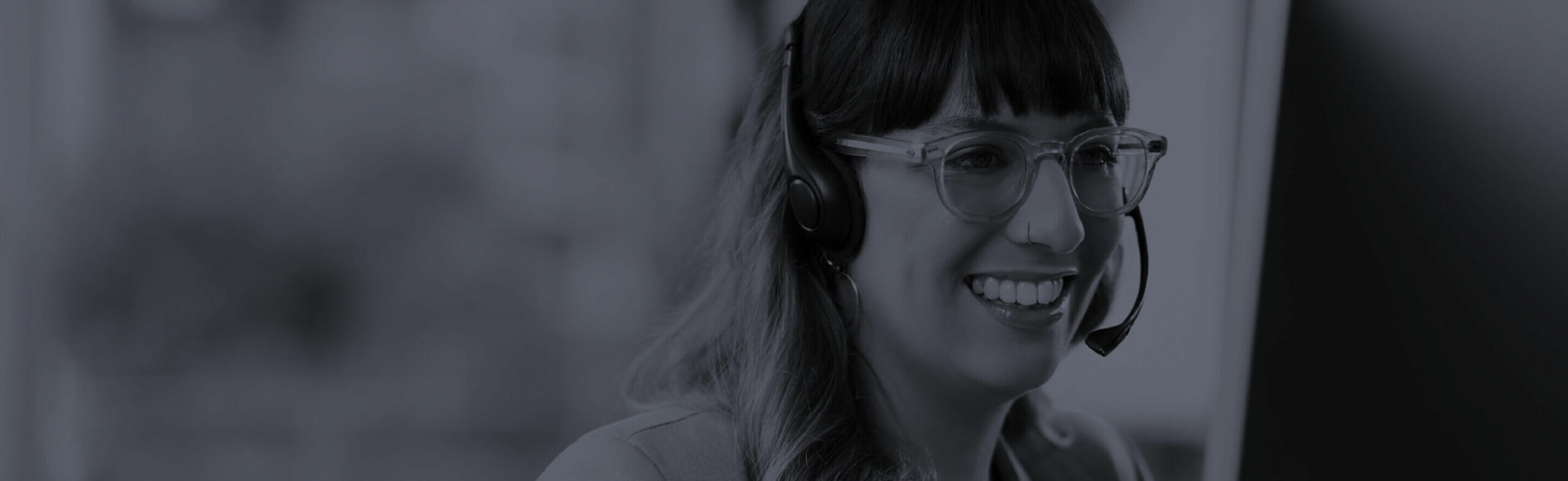Customer support representative smiling while wearing a headset
A woman wearing glasses and a headset smiles while looking at a computer screen, working in a professional office or support center.