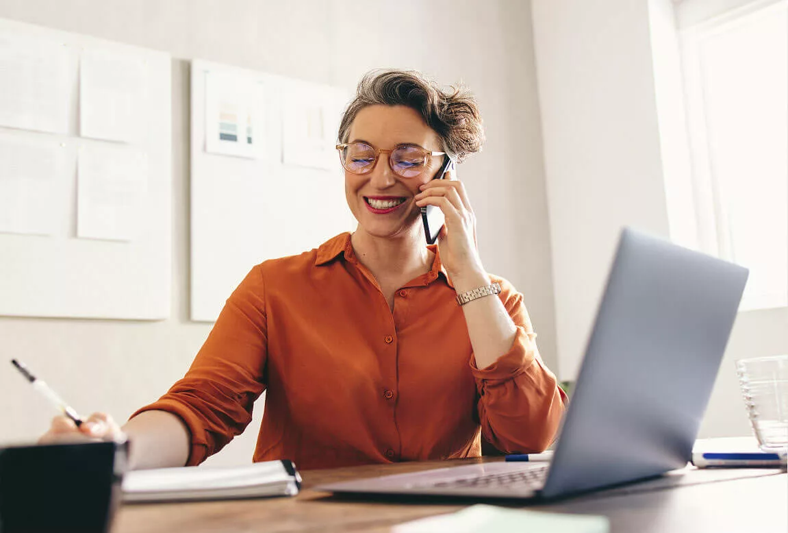 Professional woman taking a call while working at her desk
A smiling woman wearing glasses talks on a smartphone while writing notes beside a laptop in a bright office workspace.